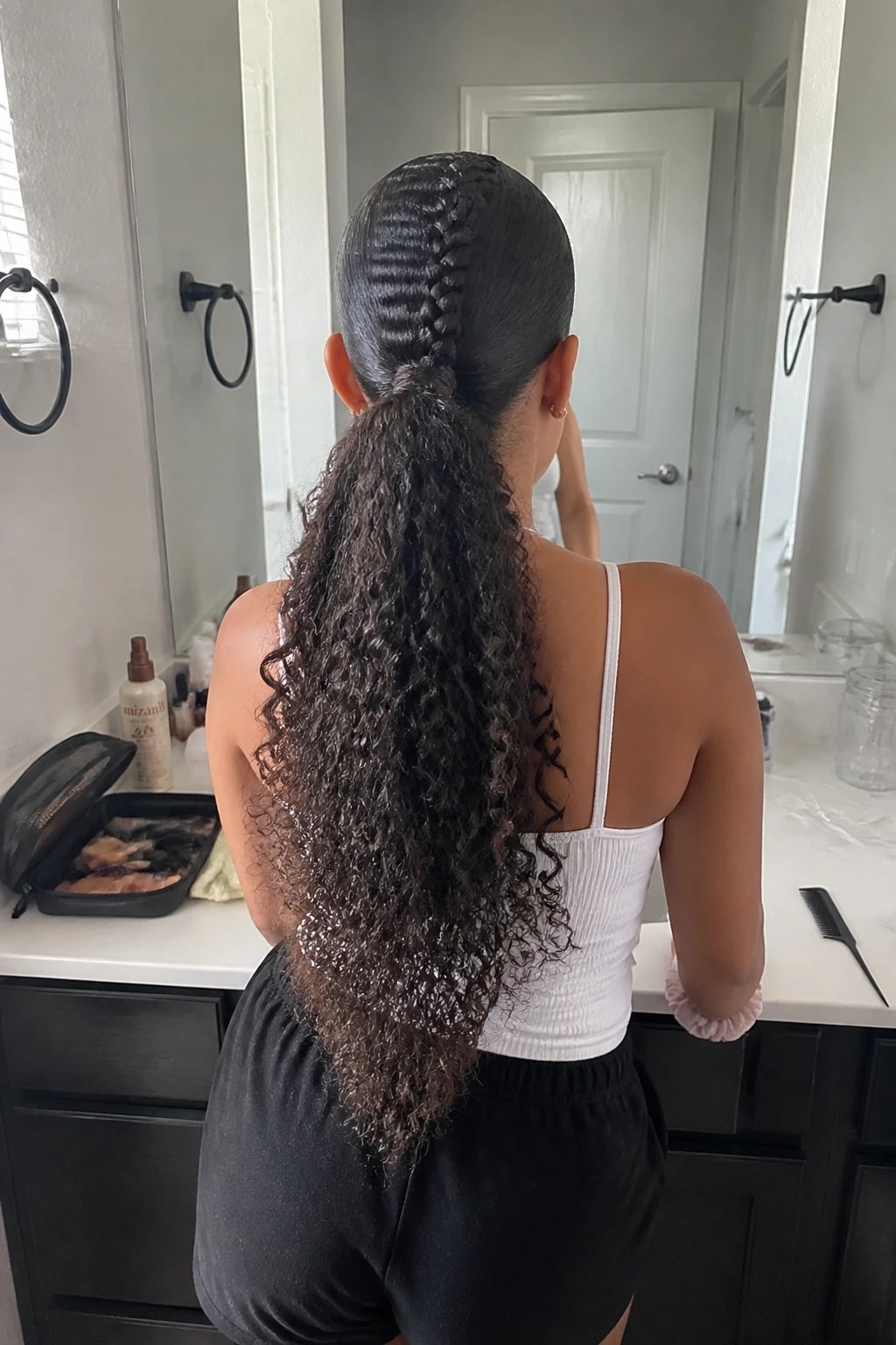 Photo d'une femme portant une queue de cheval mohawk bouclée avec une seule tresse centrale se prolongeant en une queue de cheval haute et volumineuse, les côtés soigneusement lissés, vue de l'arrière de la tête mettant en valeur la base tressée et les boucles en cascade, debout devant un miroir de salle de bain, lumière naturelle, contenant un petit détail du quotidien comme un peigne à queue à côté d'une trousse de maquillage ouverte, un flacon de rafraîchisseur de boucles à proximité et un chouchou en soie à son poignet, moment réaliste de préparation, qualité photo iPhone de dernière génération.