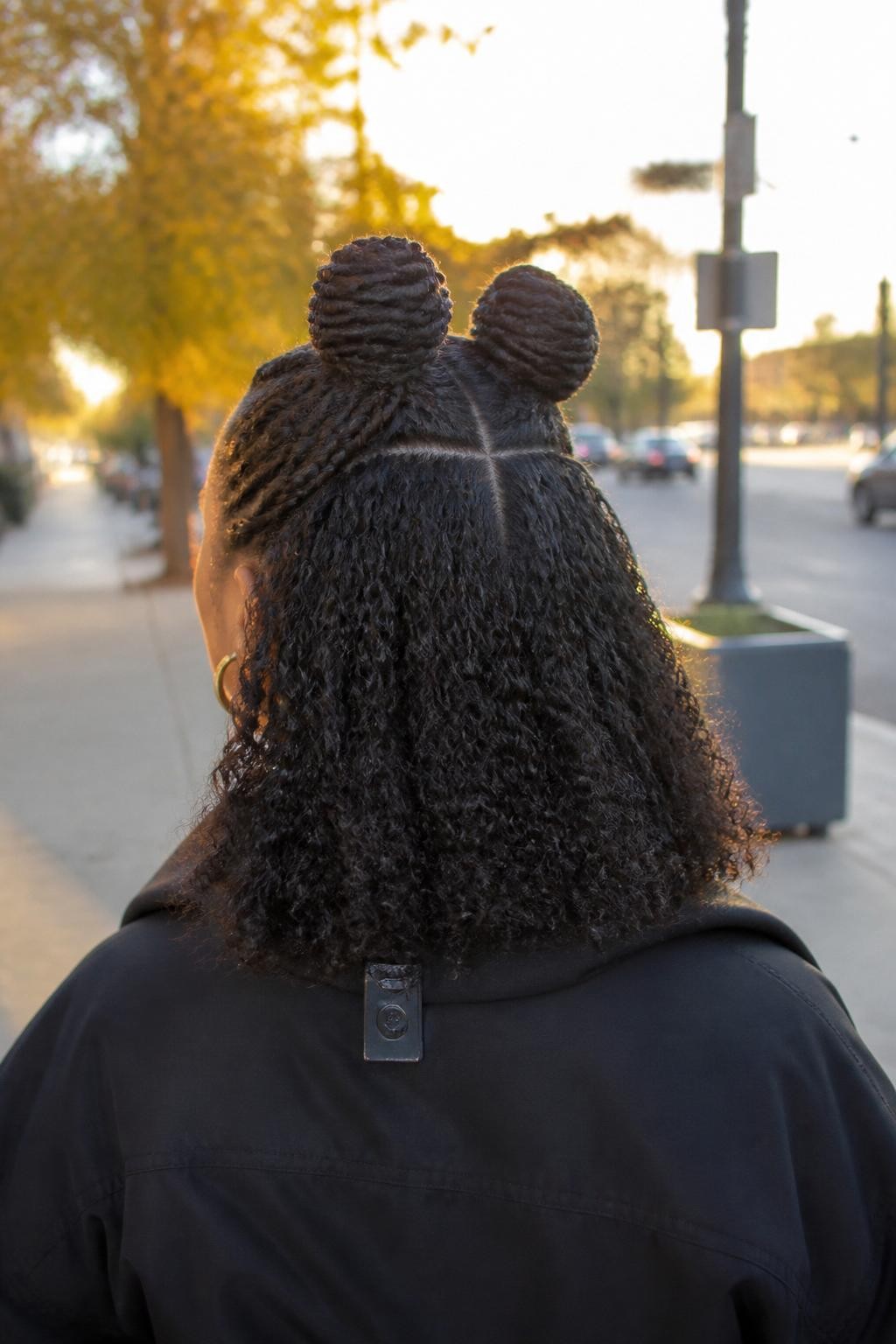 Image de référence : Photo d’une femme noire portant deux petits chignons tressés mi-hauts, avec des boucles définies mi-longues laissées libres, vue de l’arrière de la tête montrant la raie au milieu et les bases tressées, arrière-plan de rue légèrement flou avec feuillage d’automne, lumière dorée du crépuscule, pince à cheveux attachée au col de sa veste et petites boucles d’oreilles créoles visibles, photo prise sur le vif, texture de boucle réaliste, qualité photo iPhone de dernière génération.