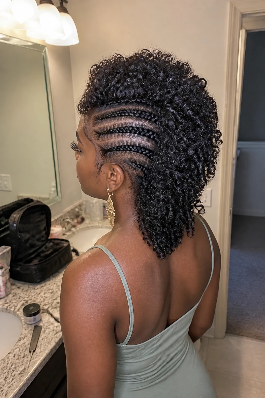 Image d'illustration : Photo d'une femme noire portant une crête iroquoise bouclée avec quatre tresses africaines soignées de chaque côté et une rangée complète de boucles naturelles et souples au centre, vue de trois quarts arrière mettant en valeur le motif des tresses et la texture des boucles, debout devant un miroir de salle de bain, éclairage intérieur doux, contenant un petit détail réaliste comme du gel pour les bords et un peigne à queue sur le comptoir, une trousse de maquillage à proximité et des boucles d'oreilles imposantes qui dépassent, moment réaliste de préparation, qualité photo iPhone de dernière génération.