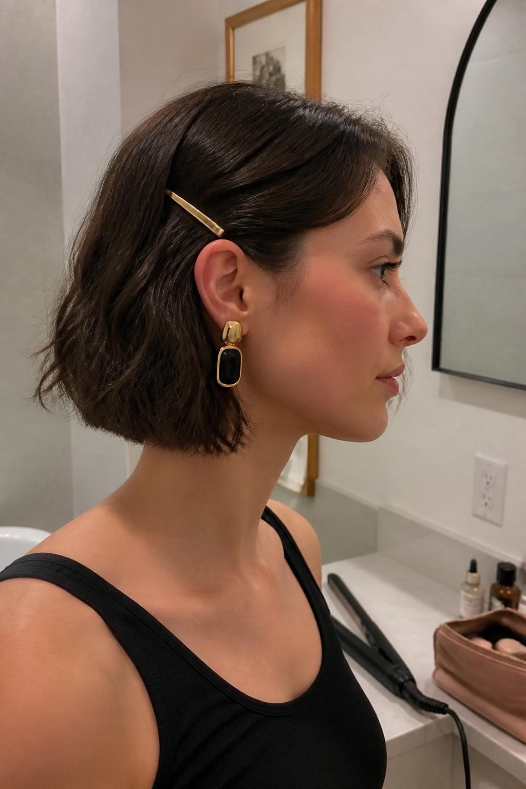 Image suggérée : Photo d’une femme portant un carré court et ondulé arrivant au menton, avec des ondulations souples réalisées au fer à lisser et une mèche relevée derrière l’oreille par une fine barrette dorée, vue de profil, miroir de salle de bain, éclairage intérieur doux, fer à lisser sur le comptoir et trousse de maquillage à proximité, boucle d’oreille imposante visible du côté relevé, texture naturelle irrégulière, qualité photo iPhone récente.
