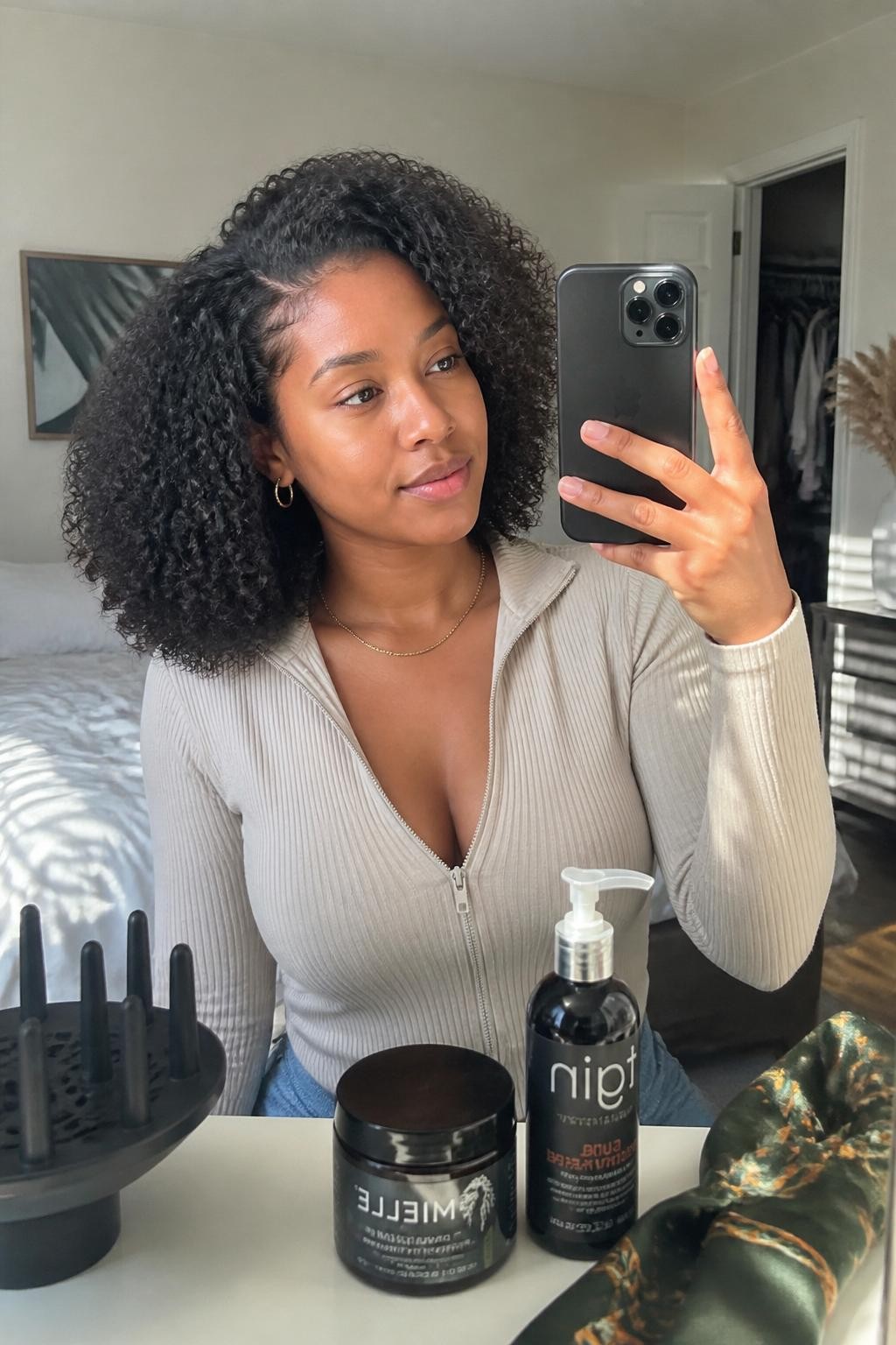 Image de référence : Photo d’une femme noire arborant une coupe au carré bien définie, mi-longue, avec des boucles souples, une raie sur le côté et des racines surélevées. Selfie pris sur le vif devant un miroir, dans une chambre baignée de soleil, avec lumière naturelle, diffuseur et crème pour boucles visibles sur la commode, foulard en satin négligemment posé à proximité et placard légèrement ouvert derrière elle. Routine capillaire naturelle réaliste, qualité photo prise avec un iPhone récent.