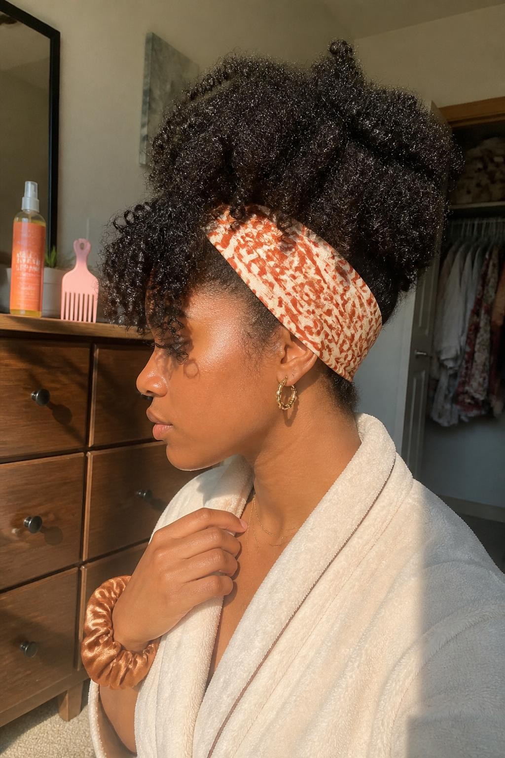 Image suggérée : Photo d’une femme noire portant une coiffure ananas bouclée haute avec des boucles bien définies retombant vers l’avant et un large bandeau imprimé rouille et crème, vue de profil spontanée, chambre baignée de soleil, lumière naturelle chaude provenant d’une fenêtre, flacon de rafraîchisseur de boucles et peigne à cheveux sur la commode, chouchou en soie autour de son poignet, peignoir confortable et placard légèrement ouvert en arrière-plan, qualité photo iPhone de dernière génération.